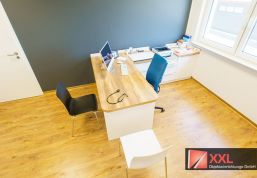 Behandlungszimmer mit ergonomischem Arbeitsplatz, schwarzem Stuhl und medizinischem Zubehör auf dem Schreibtisch.