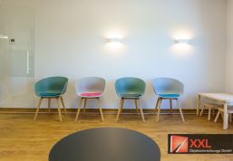Der Sitzbereich kombiniert modernes Design mit Funktionalität. Vier farblich akzentuierte Stühle stehen vor einer hellen Wand mit minimalistischer Beleuchtung.