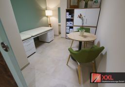 Ein Beratungsraum mit einer Untersuchungsliege in hellem Cremeweiß, kombiniert mit grünen Stühlen und einem runden Holztisch. Ein Schrank mit medizinischem Equipment und eine dezente Beleuchtung sorgen für eine angenehme Atmosphäre.