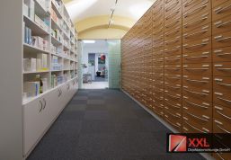 Ein Blick in das großzügige Medikamentenlager mit hohen, weißen Regalen und einem riesigen Apothekerschrank mit zahlreichen Schubladen aus Holz. Die moderne Architektur mit Gewölbedecken und Glaswänden unterstreicht die professionelle Atmosphäre.