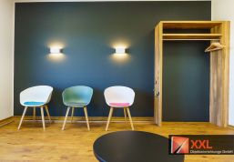 Ein stilvoller Garderobenbereich aus hellem Holz mit bunten Haken bietet Platz für Jacken und Taschen. Drei moderne Sitzschalenstühle setzen farbliche Akzente vor der dunkel abgesetzten Wand und schaffen ein harmonisches Gesamtbild.