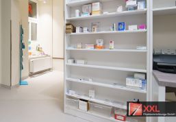 Ein weiteres Regal im hinteren Bereich der Apotheke, das mit wichtigen Medikamenten bestückt ist. Die moderne weiße Farbgestaltung sorgt für eine saubere und übersichtliche Struktur.