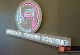 Modernes LED-beleuchtetes Praxislogo mit den Initialen „HR“ sowie dem Namen „Helene & Dr. Rüdiger Soukup“ auf einer strukturierten Betonwand – ein stilvoller Blickfang im Empfangsbereich.